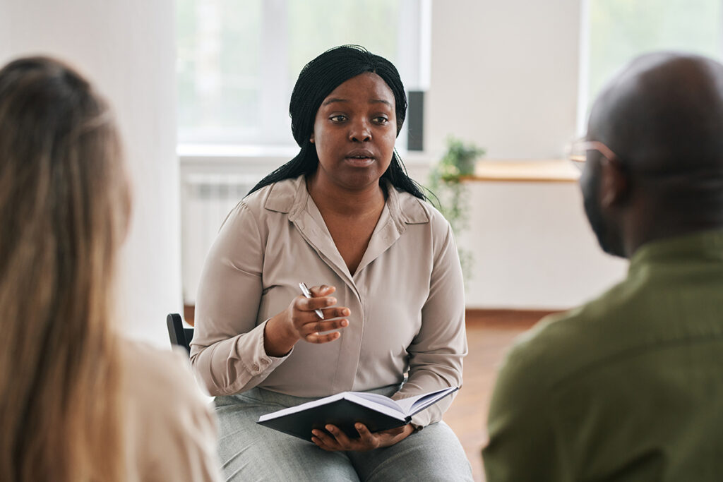 Young confident black woman with notebook and pen consulting intercultural couple sitting in front of her during session